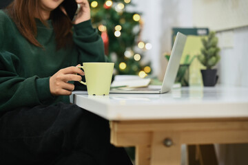 Unrecognizable young woman holding cup of coffee and talking on mobile phone in living room decorated for Christmas