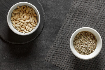Sunflower and pumpkin seeds in bowls on dark slate stone plate and grey tablecloth, top view
