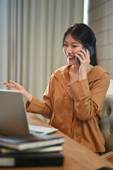 Smiling Asian female employee having pleasant phone conversation and using laptop at workplace