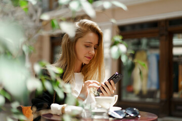 Beautiful  blond woman in stylish   casual business outfit  sitting in cafe   in  beautiful european city.  Using mobyle phone.