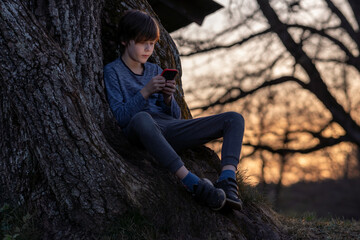 Portrait of teenage boy sitting on roots of large tree and enthusiastically watching something on...