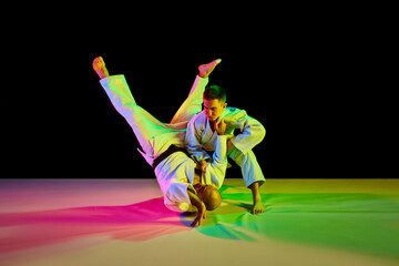 Two Caucasian karate fighters in white kimono with black belts performing skills in action in neon light isolated black. Concept of combat sport.