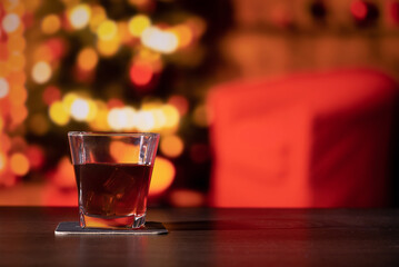 Glass of whiskey on the bar in front of the blur image Christmas home room with tree and festive bokeh lighting, blurred holiday background