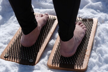 A woman practicing yoga therapy stands on a board with nails against a background of snow.