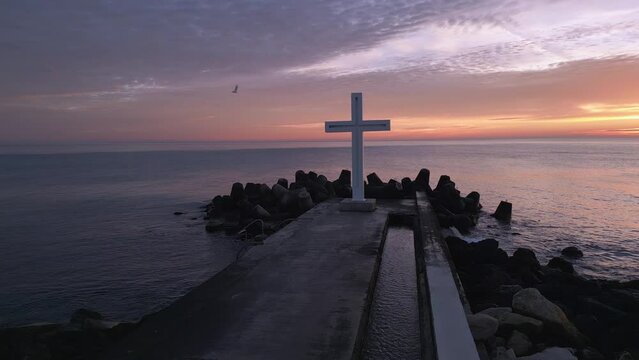 A flight around a Christian Holy cross early in the morning at sunrise. The large cross stands on the edge of a breakwater on the sea coast