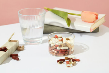Different types of pills are contained in a glassware, a weekly pill organizer, a glass of water and a tulip flower is placed on the notebook. White table and pink background. Advertising.