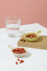 The pills are contained in small ceramic dishes with handles and a glass of water placed on a white table with a pink background. Theme of medicine.