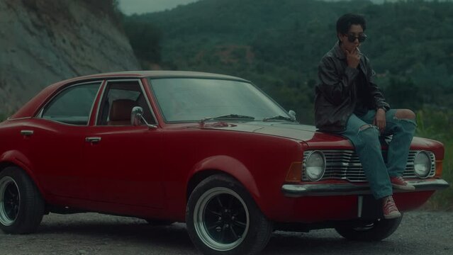 A brutal girl in a brown leather jacket sits on the hood of her vintage red car and smokes