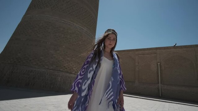 A young woman in a white dress and a long oriental robe dances in the sunshine next to an ancient minaret