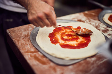 The pizza maker puts tomato sauce on the pizza dough, preparing it on the pizza shovel.