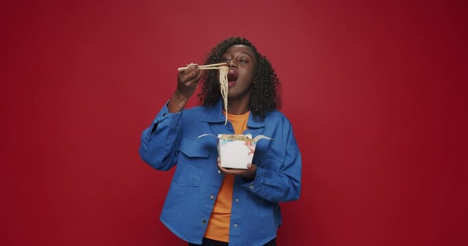 African american woman looking at camera and smiling, preparing to eat noodles, seafood and vegetables with chopsticks on isolated red background. Asia street food. Boxed fast food ready for delivery.