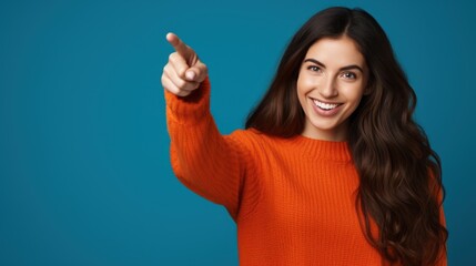 Fototapeta premium Portrait of a beautiful girl with long dark hair on a blue background wearing an orange sweatshirt, finger pointing in a side view