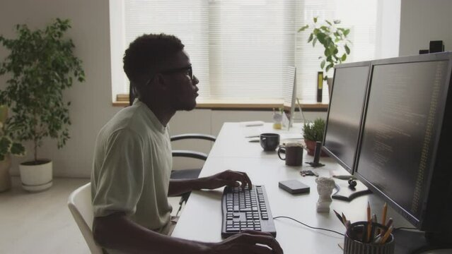 Side view waist up of young African American male geek wearing basic t-shirt and eyeglasses sitting in front of two monitors and typing program code while working in minimalist open space office