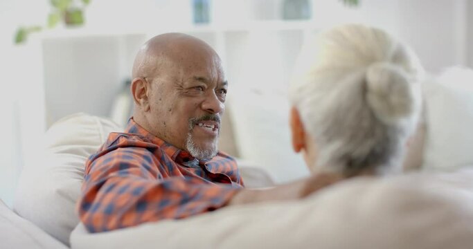 Happy senior biracial couple sitting on couch and talking at home, slow motion