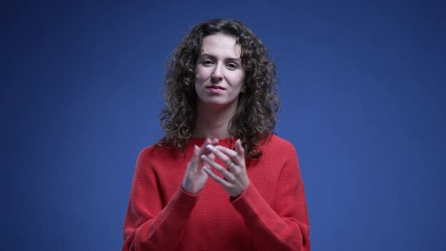 Young woman applauding with hands while looking at camera with neutral expression, standing on blue background