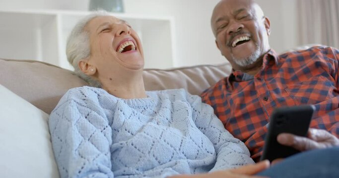 Happy senior biracial couple sitting on couch and using smartphone at home, slow motion