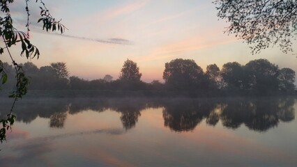 On an early spring morning, the sun, not yet above the trees on the far side of the river, colors the sky with feathery clouds in pink tones. The trees are reflected in the water, over which a mist