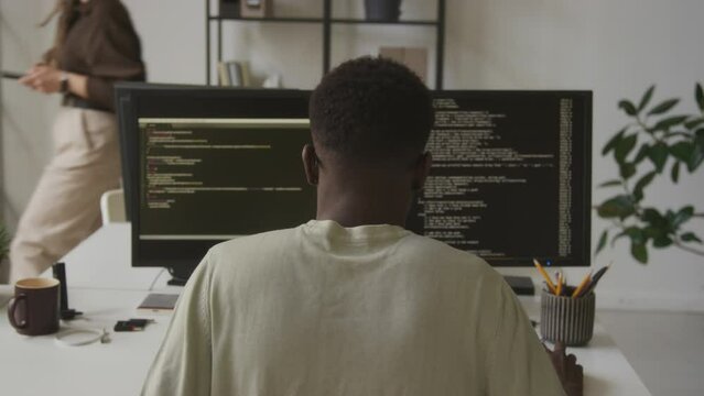 Back view of African American software engineer program coding while sitting in front of two monitors at office desk in minimalist open space office