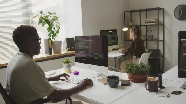 Medium shot of multiracial couple of software engineers writing programming codes on computers while working together in minimalist open space office