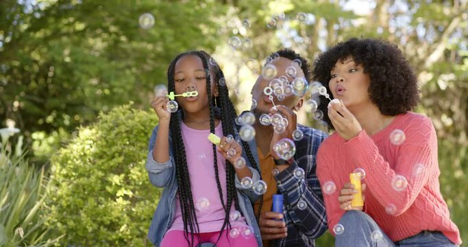 Happy african american parents with daughter blowing bubbles in garden at home, slow motion