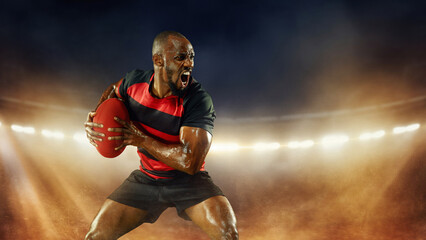 Emotional and motivated African-American young man, rugby player in motions over dark stadium with flashlight and smoke. Concept of professional sport, competition, motivation, game, championship
