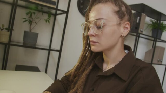 Handheld tilting shot of young focused Caucasian female software engineer in eyeglasses looking at computer monitor and typing on wireless keyboard while working in modern programmers office