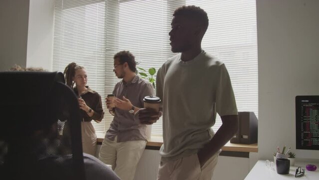 Medium shot of diverse team of programmers having coffee together at their workplaces in open space office