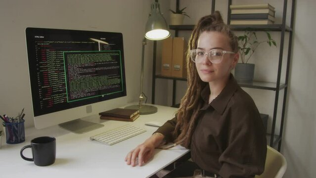Waist up portrait of young Caucasian female programmer with long dreadlocks smiling at camera while sitting at white office desk beside black computer monitor with html code on display