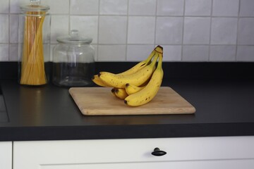 A portrait of a bunch of yellow bananas lying on a wooden cutting board on a black kitchen countertop. The delicious energizing food is ready to eat and ideal before sport.