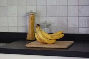 A portrait of a bunch of yellow bananas lying on a wooden cutting board on a black kitchen countertop. The delicious energizing food is ready to eat and ideal before sport.