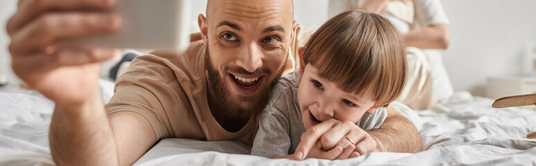 joyous bearded father taking selfies with his little son while lying on bed, family concept, banner