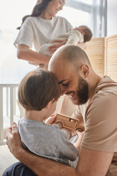 vertical focused shot of father playing with his son with his blurred wife with newborn on backdrop