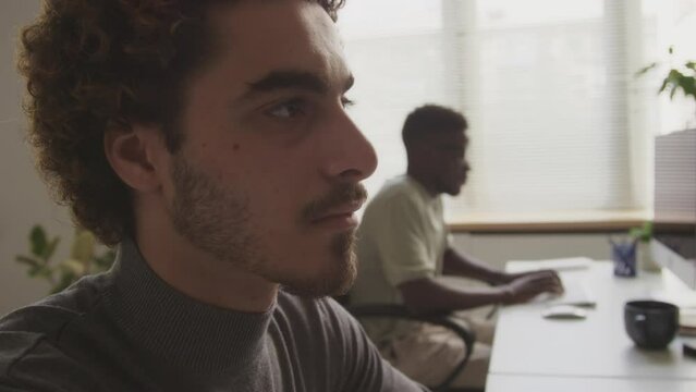 Handheld tilting up shot of young curly haired Biracial male software engineer looking at computer monitor while typing program code on wireless keyboard, working with colleague in office