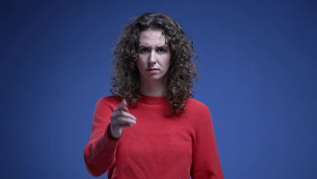 Young woman saying NO to camera by pointing and waving finger. 20s female person making cross with arms rejecting offer or behavior standing on blue backdrop