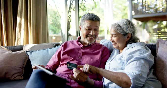 Happy diverse senior couple sitting on couch and using tablet for online shopping at home