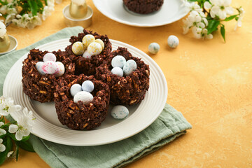 Easter chocolate nest cake with mini chocolate candy eggs with blossoming cherry or apple flowers on yellow background table. Creative recipe for Easter table with holiday decorations. Top view.