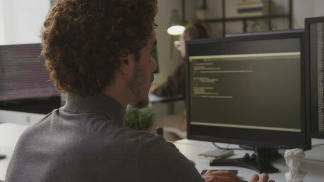 Back view shot of Biracial male programmer with beard and curly hair writing program code in computer on keyboard sitting in front of two monitors at desk in modern office