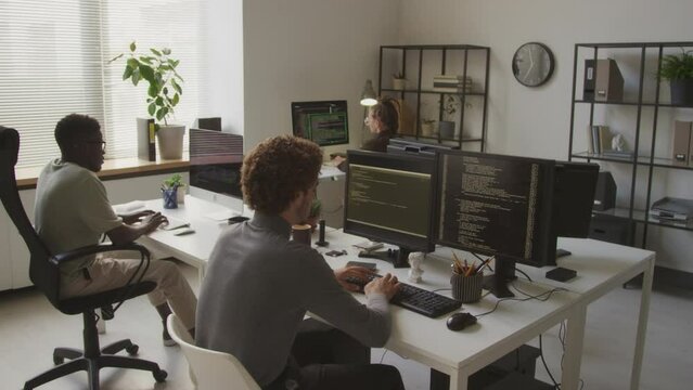 Back view of multiethnic programmers team of three writing program codes on desktop computers while working together in contemporary minimalist open space office