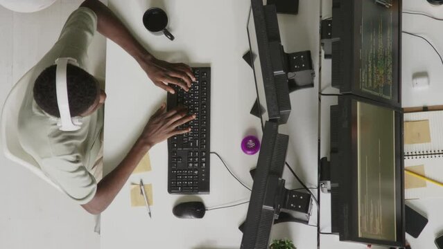 Top view stan shot of young African American male programmer in wireless headphones writing program code on keyboard while sitting in front of two monitors at office desk
