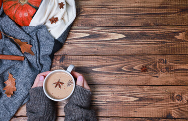 Cropped shot of female hands holding fresh coffee mug, latte, spicy cortado, bright orange pumpkin wrapped in woolen scarf, dry oak leaves, spices on rustic wooden table. Autumn background