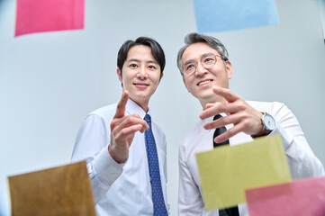 Two businessmen are standing on the glass wall of the office, looking at the attached paper and...