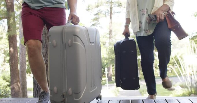 Diverse senior couple walking with luggage to a house in sunny outdoors