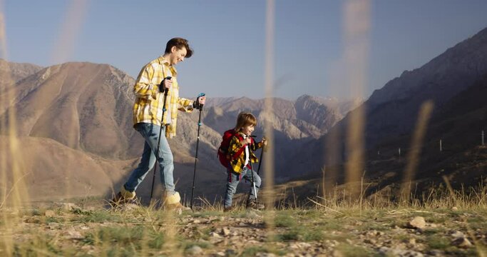Mother and son conquer mountain, trekking poles backpacks. Mother and son traverse rocky path. Confidence, walk on dry grass. Small steps towards great goal, importance of support. Ecotourism, family