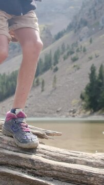 A young adventurer strolls along a fallen tree by a mountain lake in a U.S. national park. Focus on legs and boots, showcasing a childhood immersed in nature. The vertical video captures the essence