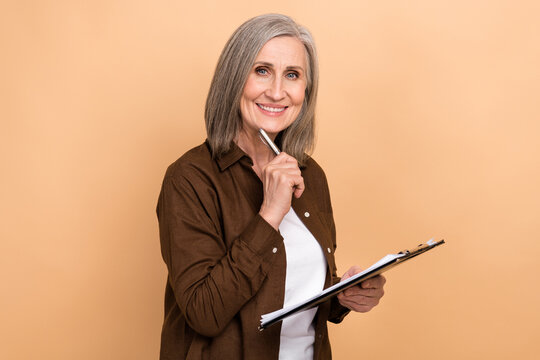 Photo of positive aged person hold clipboard pen touch chin contemplate isolated on beige color background