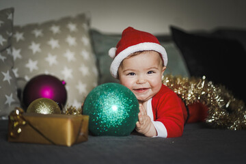 happy smiling baby boy in red santa claus costume with christmas hat playing with christmas balls at home on grey background
