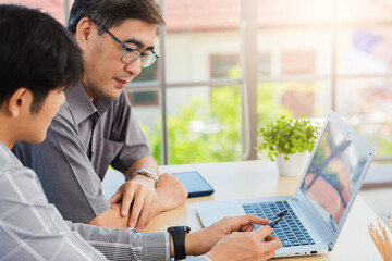 Asian young man teaching senior father how to use credit card for shopping online with laptop computer in the living room, adult son holding bank card helping Dad to purchases web online application