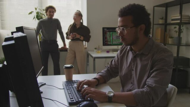 Waist up shot o Biracial male programmer working on computer in office while his multiethnic colleagues talking by windowsill in background
