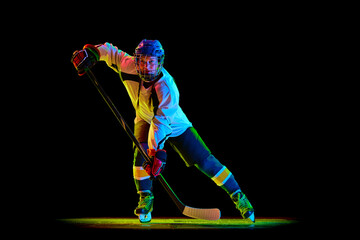 Young girl, hockey player in uniform and helmet training, standing with stick against black studio background in neon light. Concept of professional sport, competition, game, action, hobby