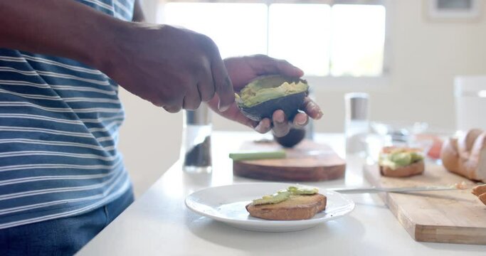 African american man putting avocado on toast in sunny kitchen, slow motion